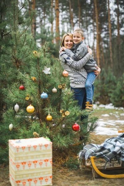 Güzel, genç bir anne oğlunu açık havada, Noel ağacının yanında tutuyor. Ağacın altındaki tahta kızak ve hediye kutuları. — Stok fotoğraf