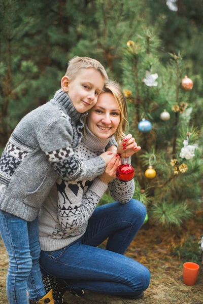 Famille, jeune mère et petit fils, portant des pulls tricotés, décorant sapin dans la forêt ou le parc. Décorations colorées, cadeaux de Noël — Photo