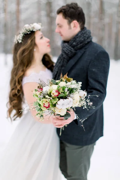 Zimní svatba, stylový mladý pár, nevěsta a ženich, v lese, něžná objetí. Nevěsta s dlouhými vlasy v bavlněném věnci na hlavě drží zimní svatební kytici. Zaměření na kytici — Stock fotografie