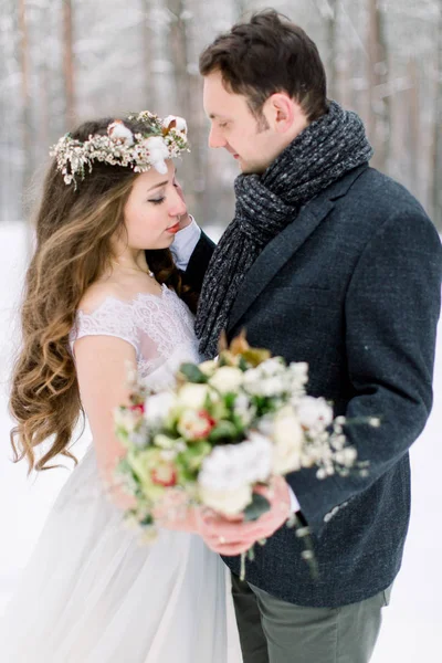 Свадебная пара зимой в снежном лесу. Красивый жених обнимает свою невесту и трогает венок на ее голове — стоковое фото