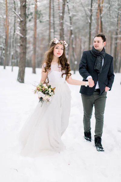 Wedding in winter forest, bride in elegant dress with wreath on head and flower bouquet and groom walking at winter wedding day — Stock Photo, Image