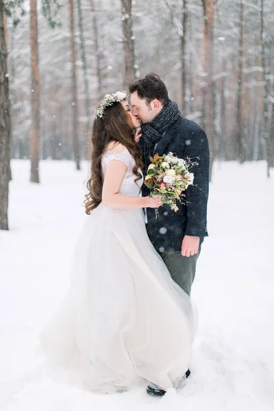 Winter wedding couple, bride and groom hugging and kissing in the snowy forest at their wedding walk