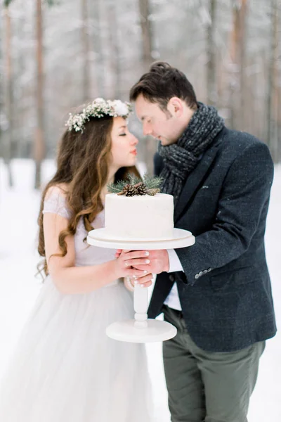 Vackra par brud och brudgum som håller vinter bröllop kaka, stående utomhus i vinter snöig skog — Stockfoto