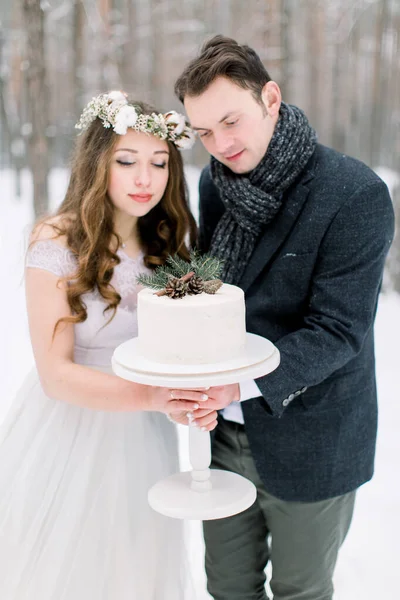 Noiva em coroa de flores e noivo elegante segurando bolo decorado de casamento de inverno. Casamento de inverno na floresta nevada — Fotografia de Stock