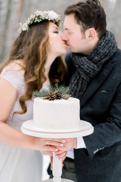 A wedding couple holds a beautiful white wedding cake, decorated with pine branch, cones and cinnamone, standing over the background of winter forest — Stock Photo, Image