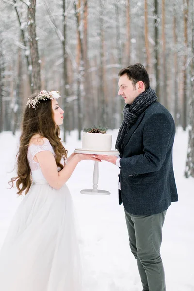 Atraktivní nevěsta s dlouhými kudrnatými vlasy stojící spolu se svým ženichem ve stylové bundě, držící svatební dort v zimě zasněžený les — Stock fotografie