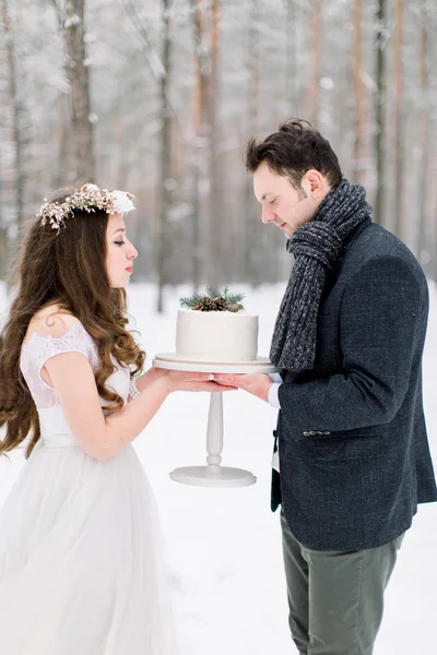 Noiva atraente com cabelos longos encaracolados em pé, juntamente com seu noivo na jaqueta elegante, segurando bolo de casamento na floresta nevada de inverno — Fotografia de Stock