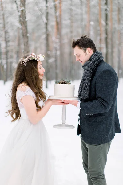 Attraktiv brud med långt lockigt hår står tillsammans med sin brudgum i snygg jacka, håller bröllop tårta på vintern snöig skog — Stockfoto