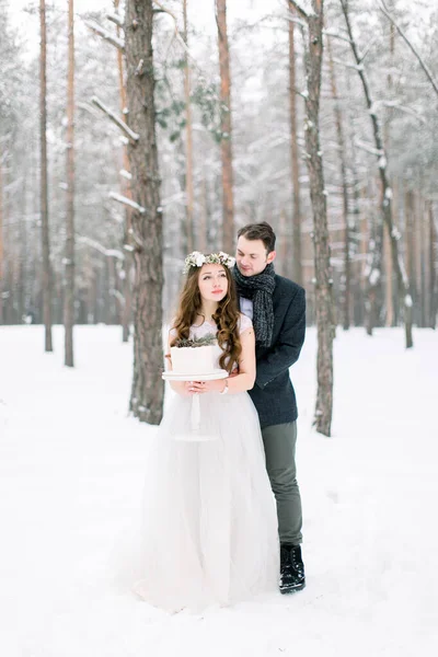 Bruden och brudgummen omfamnar på en bakgrund av en vinterskog. Vacker kvinna brud håller dekorerad vinter tårta — Stockfoto