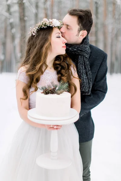 Beau couple de mariage posant avec gâteau décoré dans les mains debout à l'extérieur dans la forêt, marié embrassant sa mariée. Hiver forêt enneigée fond — Photo