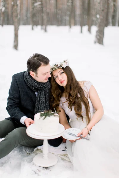 Beautiful wedding couple sitting in winter snowy forest. Decorated wedding cake