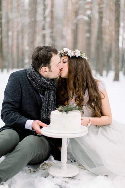 Floresta de inverno e história de amor do casamento. Bolo de inverno decorado, e belo casal sentado e beijando, floresta de inverno no fundo — Fotografia de Stock