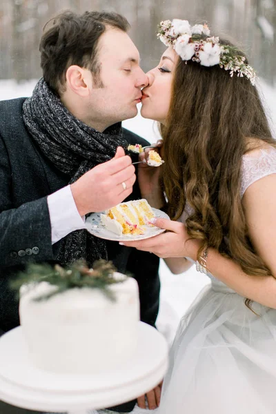 Stylish pretty bride and groom eating beautiful decorated winter wedding cake and kissing. Winter wedding in snowy forest — Stock Photo, Image