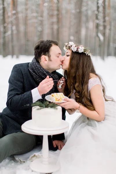 아름답고 아름다운 신부와 신랑 이 아름다운 겨울 웨딩 케이크와 키스를 먹고 있다. 눈덮인 숲 속에서의 겨울 결혼식 — 스톡 사진
