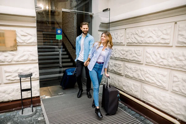 Mooie vrouw en knappe man die elkaars hand vasthouden verlaten een hotel met hun koffers. — Stockfoto