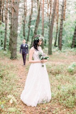 Çiçekli çelenk takmış, elinde buketle ormanda duran güzel bir gelin. Yakışıklı damat arka planda gelinine gidiyor. Kırsal düğün açık havada.