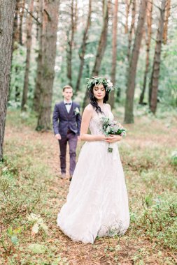 Çiçekli çelenk takmış, elinde buketle ormanda duran güzel bir gelin. Yakışıklı damat arka planda gelinine gidiyor. Kırsal düğün açık havada.