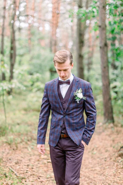 Novio guapo en traje elegante de pie sobre el fondo del bosque de pinos. Hermoso hombre, novio posando y preparándose para la ceremonia de boda rústica al aire libre — Foto de Stock