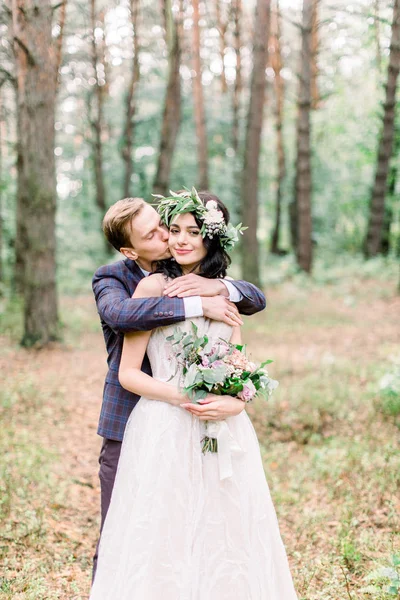 Šťastný a usměvavý svatební pár v lese. Hezký ženich balí svou krásnou nevěstu s květinovým věncem na hlavě a drží kytici — Stock fotografie