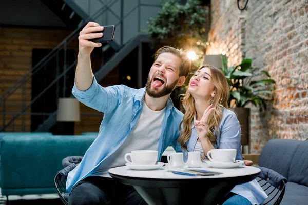 재미있는 젊은 사랑하는 부부 가 카페나 호텔에 앉아서 커피를 마시면서 스마트폰으로 셀카 사진을 찍고 있습니다 — 스톡 사진