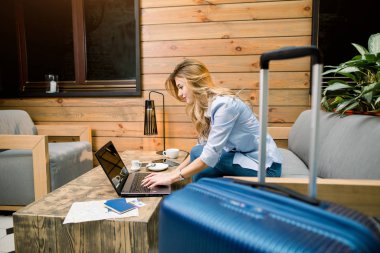 İş, seyahat konsepti. Modern otel lobisindeki kanepede otururken gülümseyen, yazan ve dizüstü bilgisayara bakan güzel bir kadının yan görüntüsü. Pasaportlar ve biletler masada, mavi bavul.