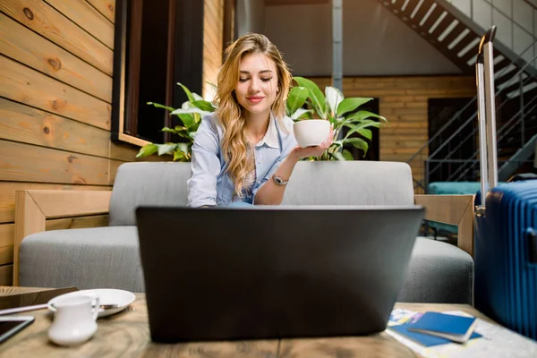 Businesswoman on trip working on laptop in lounge looking at monitor. Young pretty woman working at hotel lobby with laptop and drinking coffee — 스톡 사진