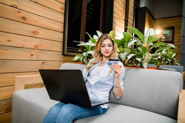 Happy young confident businesswoman holding credit card while sitting on a couch at modern office or hotel with laptop computer, online payment and booking concept — 스톡 사진