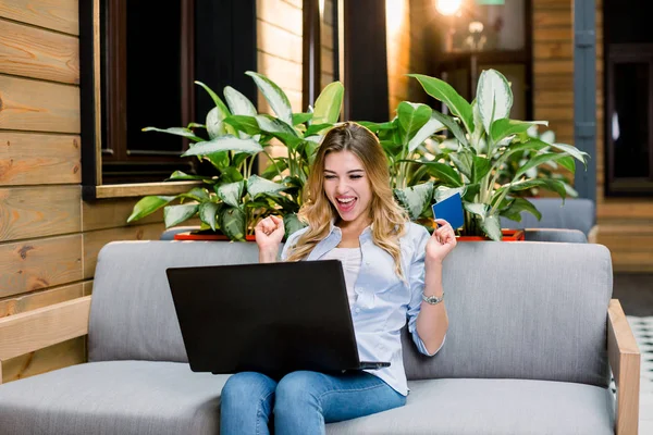 Jovem mulher loira feliz em suéter sentado no sofá com computador portátil, segurando passaporte e olhando para o laptop sobre o fundo do corredor moderno. gesto de sucesso vencedor — Fotografia de Stock