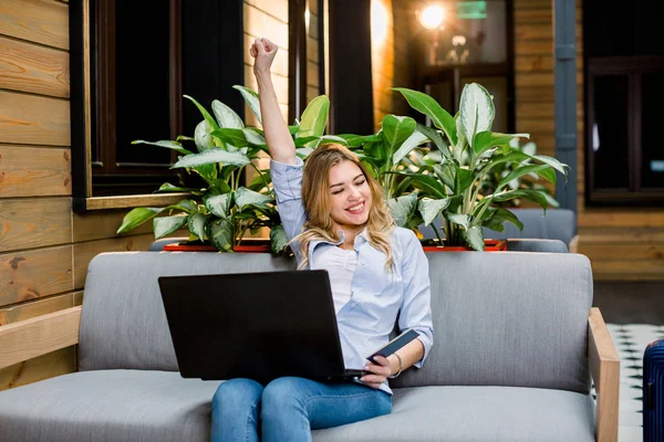 Portrait of an excited young woman in casual wear sitting on the sofa in modern loft coworking place or hotel, using laptop computer and holding passport, celebrating successful payment or booking — 스톡 사진