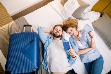 Bir çift turist manzaralı, rahat giyimli güzel kadın ve yakışıklı adam, otel odasında yatıyorlar, pasaportlarını gösteriyorlar. Seyahat, otel, rezervasyon konsepti