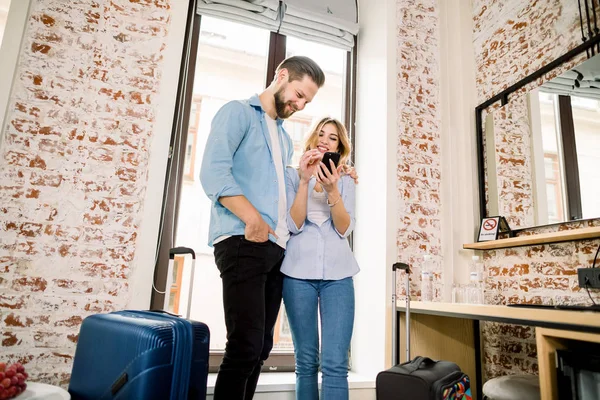 Couple of tourists with luggage ready for travel booking the tickets online using mobile phone, standing near the window in modern hotel room with brick walls — 스톡 사진