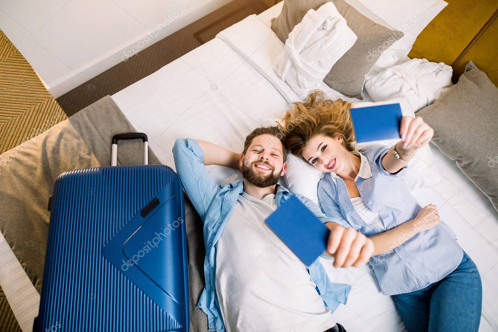 Overhead view of couple of tourists, pretty woman and handsome man in casual wear, lying on bed in hotel room, showing their passports. Travel, hotel, booking concept