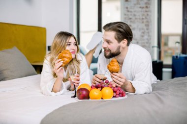 Kahvaltı, meyve, kruvasan, şampanya. Beyaz bornoz giyen bir adam ve bir kadın otel odasında kahvaltının tadını çıkarıyor.
