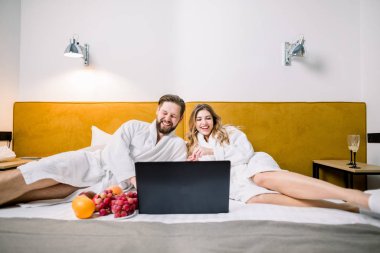 Neşeli adam ve mutlu sarışın kadın otel odasında dizüstü bilgisayara bakıyor ve film izliyor, gülümsüyor ve gülüyor.