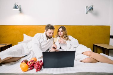 Şampanya bardaklı, dizüstü bilgisayar kullanan gülümseyen bir çift. Oteldeki yatakta içki içen çift.