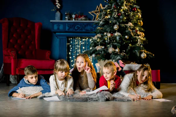Un groupe de jeunes enfants s'allongent sur une couverture de laine chaude sur le fond d'un arbre de Noël et écrivent une lettre au Père Noël — Photo