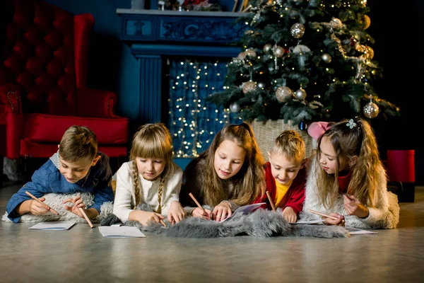 Un groupe de jeunes enfants s'allongent sur une couverture de laine chaude sur le fond d'un arbre de Noël et écrivent une lettre au Père Noël — Photo