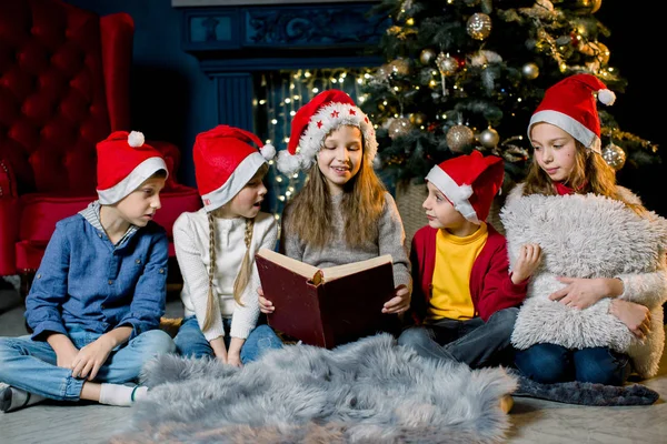 La veille de Noël, de beaux petits enfants regardent un vieux livre avec des histoires de Noël dans une chambre chaleureuse et confortable en chapeaux rouges du Nouvel An — Photo