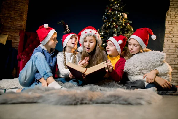 Charmante en mooie kleine kinderen in Santas hoedjes lezen kerstverhalen en lachen, op de achtergrond van een kerstboom. — Stockfoto