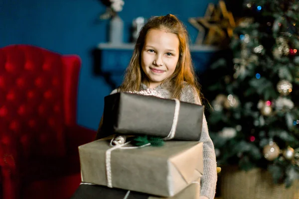 Petite belle fille heureuse tient cadeaux de Noël debout sur le fond de sapin de Noël décoré et fauteuil rouge . — Photo