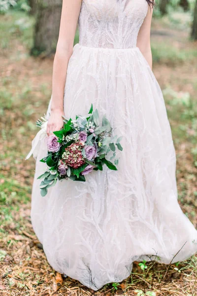 Image recadrée de jambes de belle mariée marchant dans une forêt de conifères en robe de mariée luxueuse, tenant bouquet de mariage rustique — Photo