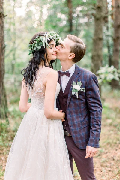 Невеста в венке на голове и элегантное платье и жених в стильном костюме ходить в лесных поцелуев. Счастливых молодоженов на свадьбе в природе, зеленый сосновый лес. Сельская свадебная пара — стоковое фото