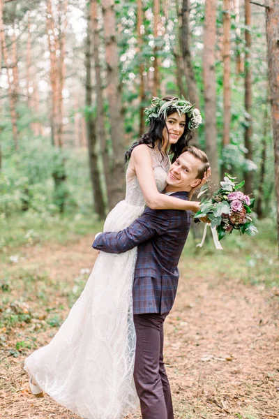 Hezký mladý muž ve stylovém obleku s brunetkou nevěstou v náručí. Romantičtí novomanželé procházejí borovým lesem. — Stock fotografie