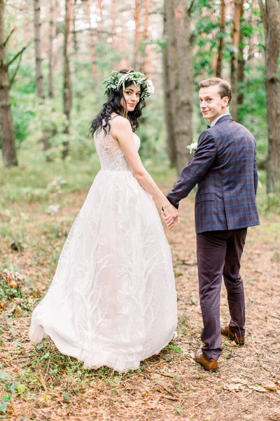 Обратный вид счастливой улыбающейся стильной невесты в цветочном венке и красивого жениха, гуляющего по лесу, развлекающегося в день своей свадьбы — стоковое фото