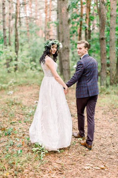 Pohled zezadu na šťastně se usmívající stylovou nevěstu v květinovém věnci a pohledného ženicha kráčejícího lesem, bavícího se v jejich svatební den — Stock fotografie