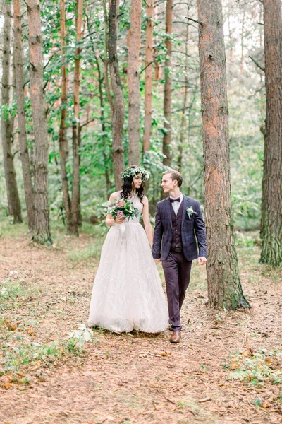 Стильная деревенская парочка молодоженов в день их свадьбы, гуляющих по лесу. Счастливая молодая невеста, элегантный жених держится за руки — стоковое фото