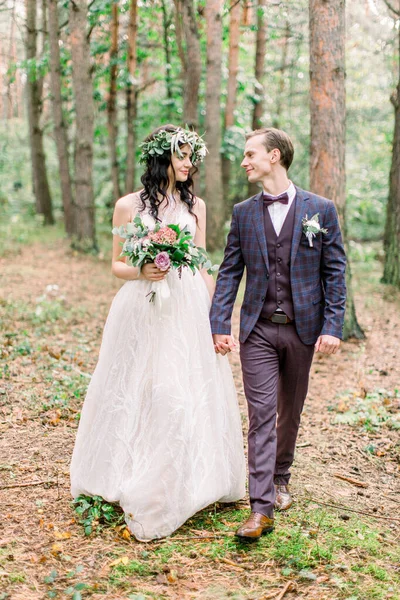 Счастливая свадебная пара в деревенском стиле, держась за руки, улыбаясь и глядя друг на друга, гуляя по лесу. Свадебная прогулка — стоковое фото