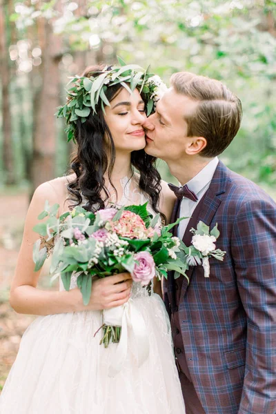 Bride in wreath on head and elegant dress and groom in stylish suit walking in forest and kissing. Rustic Wedding Couple — Stock Photo, Image