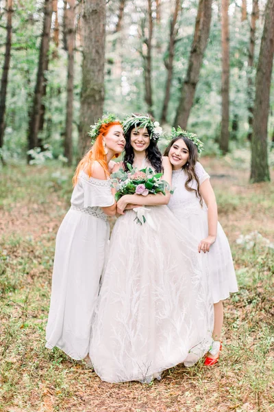 Pretty bride and two bridesmaids in white dresses smiling and looking at camera while standing on the background of pine forest. Stylish and rustic wedding. Marriage concept — 스톡 사진
