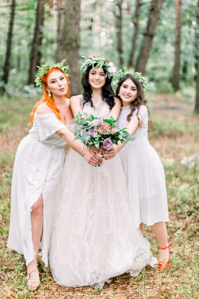 Belle mariée en robe rustique élégante et couronne tient bouquet de fleurs dans ses mains. Deux jolies demoiselles d'honneur en robes blanches et des couronnes serrent la mariée dans leurs bras. Mariage rustique dans la forêt — Photo